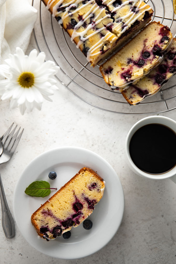 A slice of lemon blueberry loaf bread on a round white plate beside a white cup of coffee, 3 forks, a white daisy and the remaining loaf on a round cooling rack.