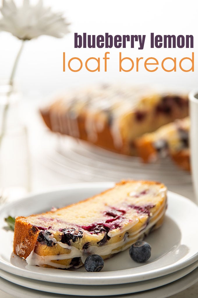 A slice of lemon blueberry bread on a round white plate beside 3 forks, a small vase with a daisy and the remaining bread on a baking rack.