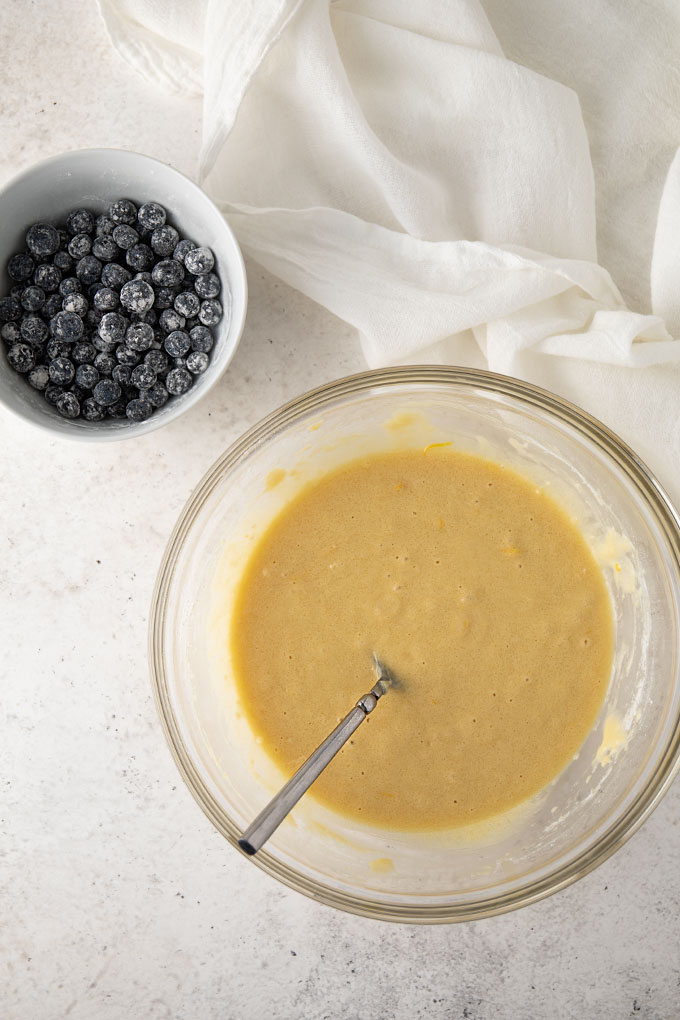 A clear glass bowl of lemon bread batter beside a white bowl of blueberries tossed in flour.