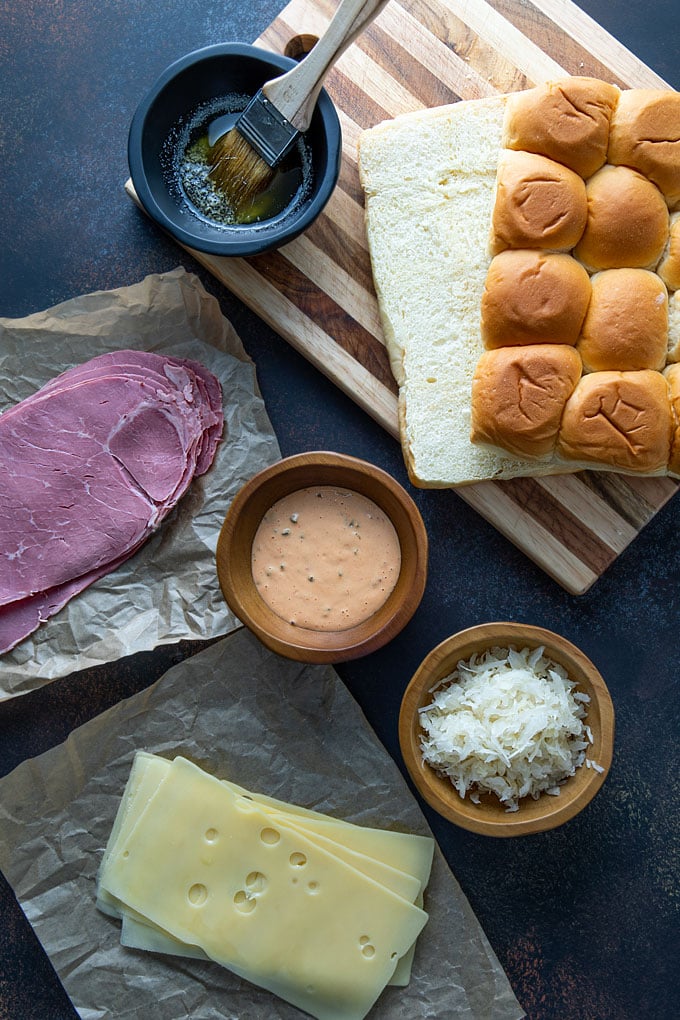 Reuben Slider Ingredients - Hawaiian sweet rolls on a wooden cutting board, a bowl with melted butter mixture, sliced corned beef, sliced Swiss cheese, thousand island dressing and sauerkraut.
