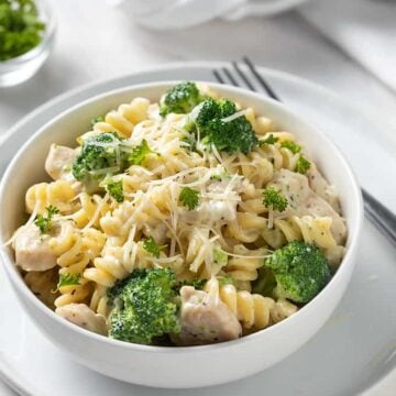 Chicken and Broccoli Alfredo in a round white bowl on a white plate with a fork on the right side.