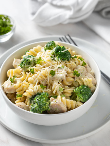 Chicken and Broccoli Alfredo in a round white bowl on a white plate with a fork on the right side.