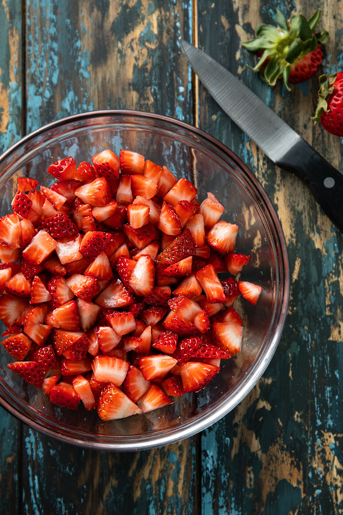 Overhead shot of a round glass bowl filled with chopped fresh strawberries beside a paring knife and a fresh strawberry.