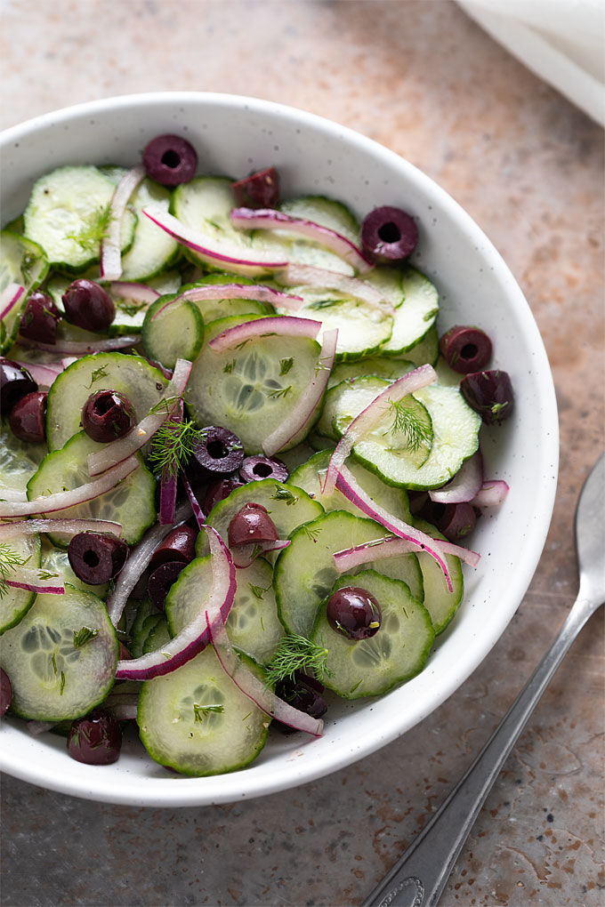 Closeup of cucumber, olive and dill salad in a round white bowl beside a serving spoon.
