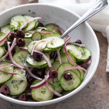 Cucumber, Olive and Dill Salad in a round white bowl with a serving spoon beside a white kitchen towel.