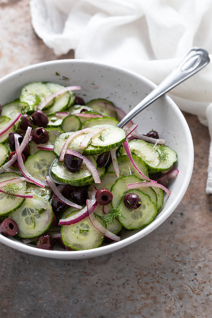 Cucumber salad in a round white bowl with a serving spoon beside a white kitchen towel.