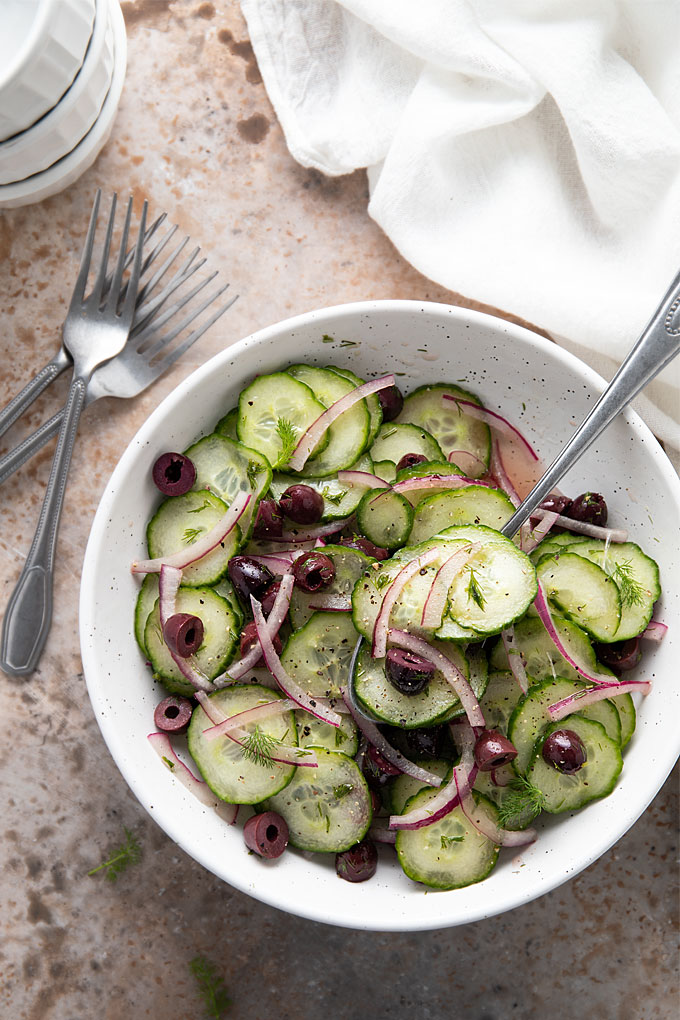 Cucumber salad in a white bowl with a serving spoon beside a white kitchen towel and 3 forks.