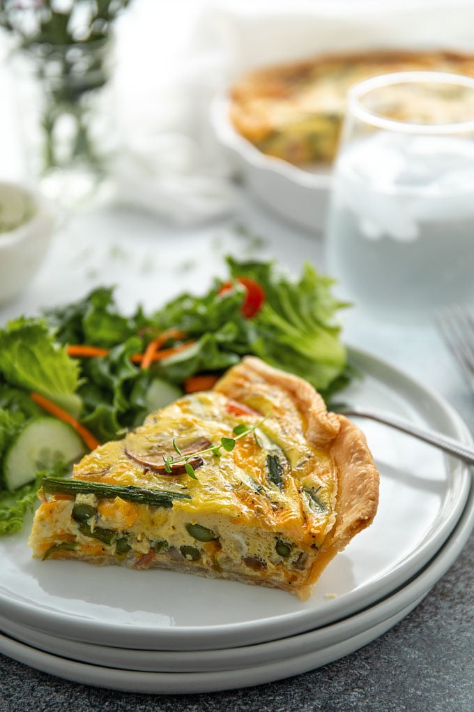 Front view of a slice of ham, asparagus and mushroom quiche on a round white plate with a garden salad.