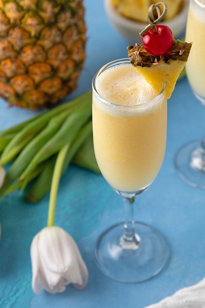 A pineapple cream mimosa garnished with a pineapple wedge beside a fresh tulip flower.