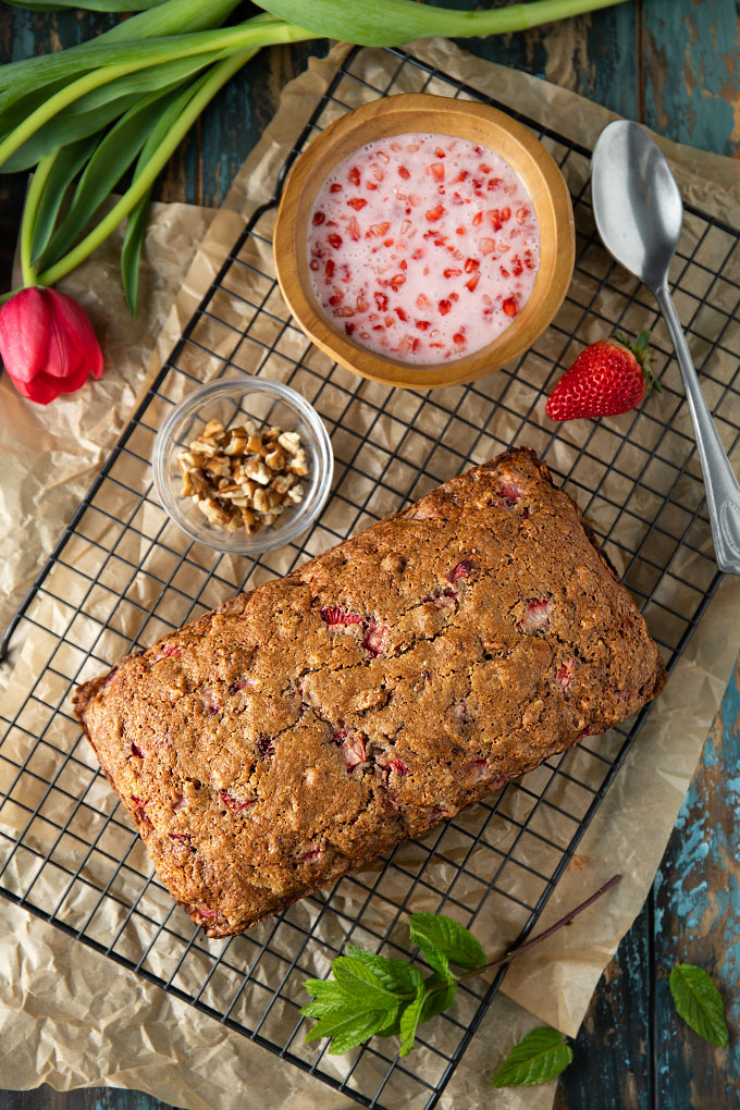 Overhead shot of a loaf of strawberry pecan bread on a cooling rack beside a bowl of chopped pecans and strawberry glaze.