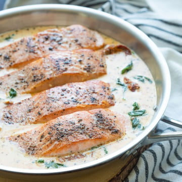 Four salmon fillets with cream sauce in a stainless skillet.