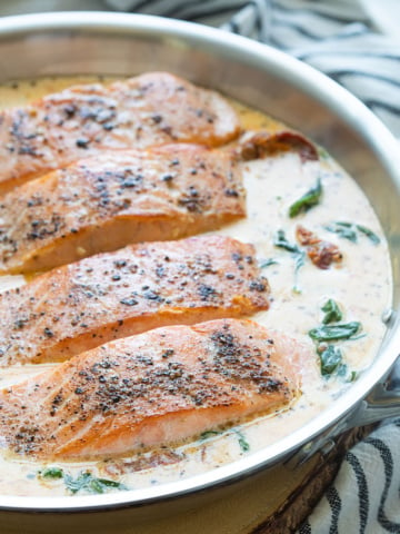 Four salmon fillets with cream sauce in a stainless skillet.