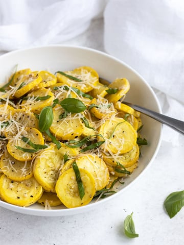 Sauteed yellow squash with fresh basil and shredded Parmesan in a round white bowl beside a white napkin.