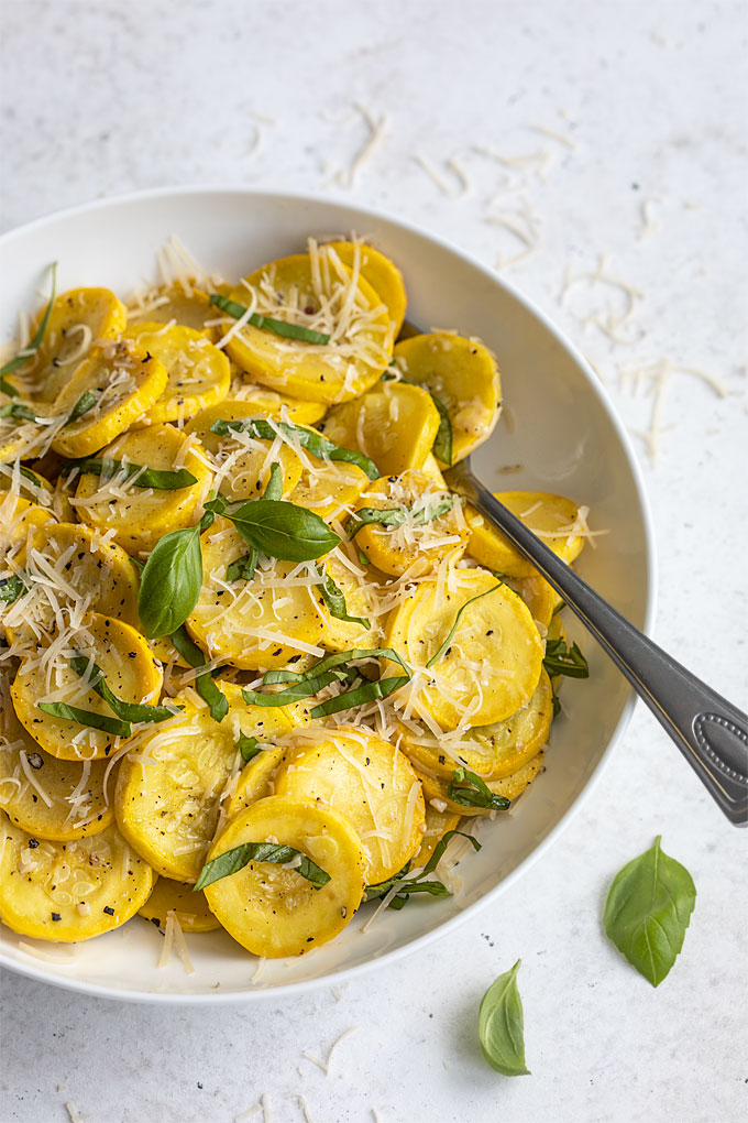 Sauteed yellow squash with fresh basil and shredded Parmesan in a round white bowl with a serving spoon.