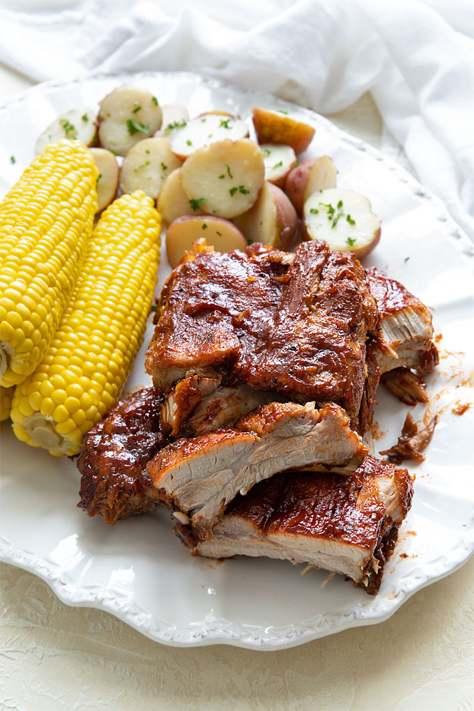 Overhead view of a rack of ribs, 3 ears of corn on the cob and baby red potatoes on a white oval serving platter