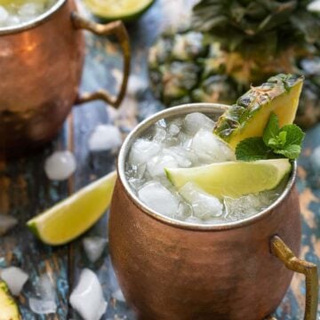 2 Moscow Mule cocktails in copper mugs garnished with fresh pineapple, lime wedges and mint sprigs