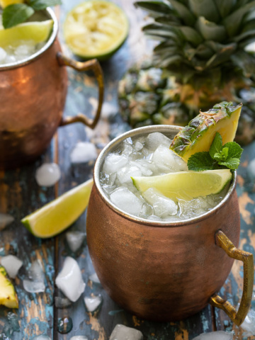 2 Moscow Mule cocktails in copper mugs garnished with fresh pineapple, lime wedges and mint sprigs
