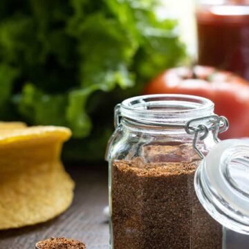 Taco seasoning in a spice jar with taco shells, lettuce and a tomato in the background
