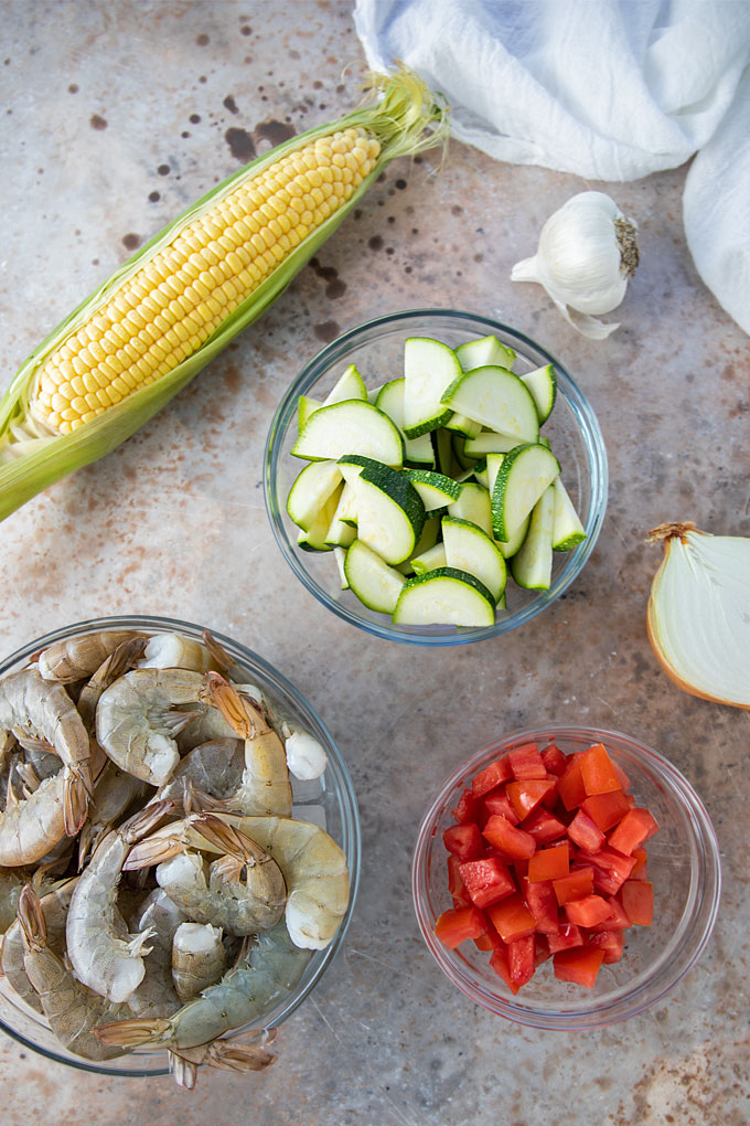 Overhead view of corn on the cob, shrimp, zucchini, tomatoes, onion and garlic.