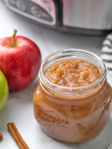 Applesauce in a mason jar beside fresh apples with a slow cooker in the background