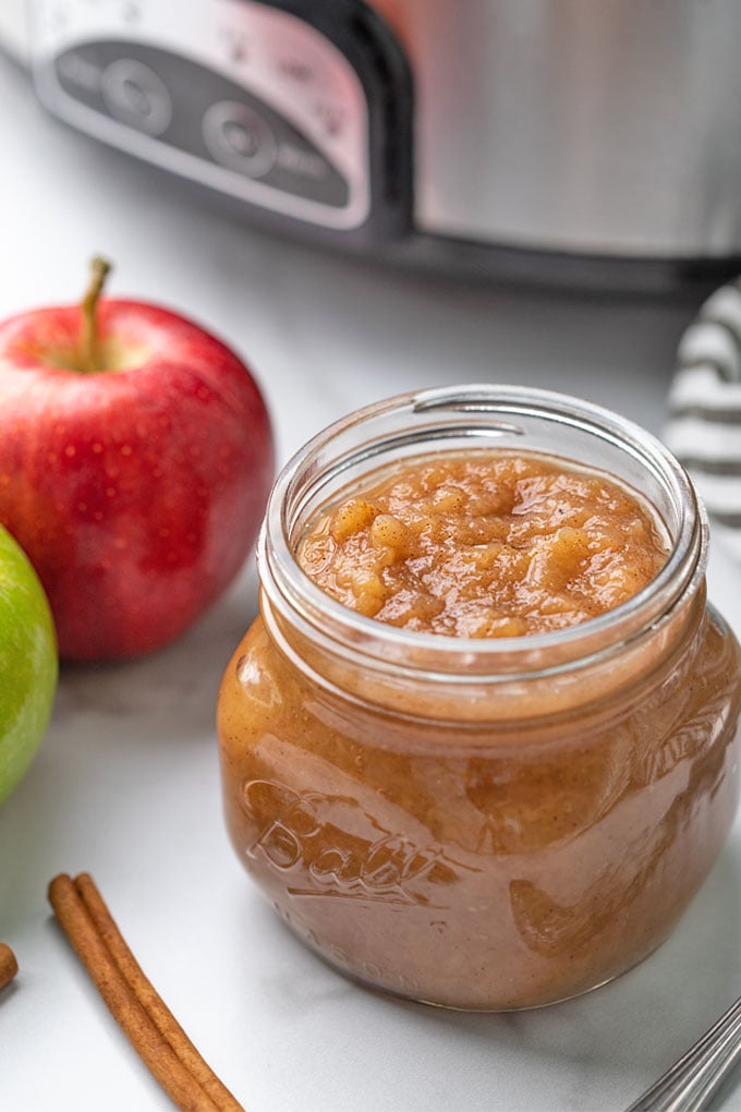 Applesauce in a jar beside fresh apples with a slow cooker in the background.