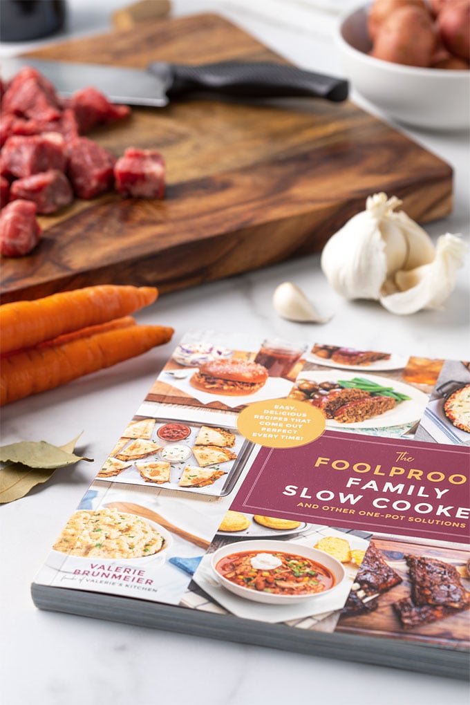 A cookbook on a marble surface with carrots, garlic and beef in the background.