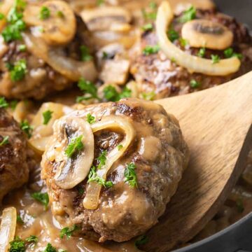 Hamburger steak patties with gravy, mushrooms and onions in a skilletwith a wooden spatula