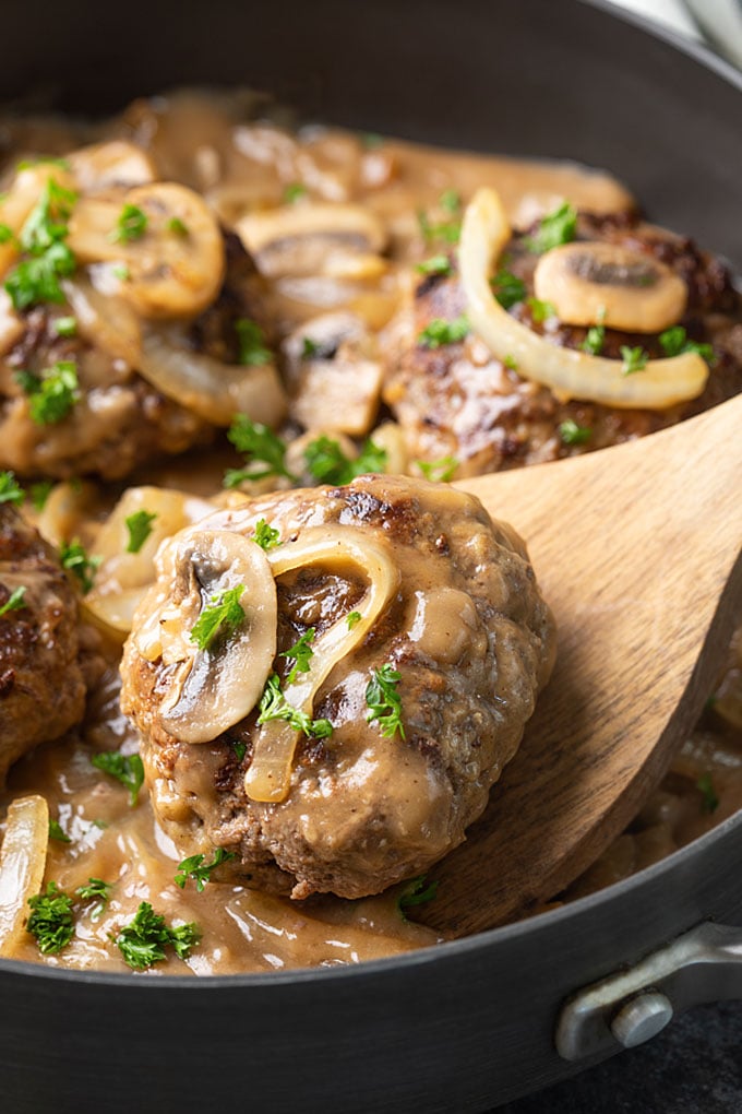 Hamburger steak patties with gravy, mushrooms and onions in a skillet with a wooden spatula.