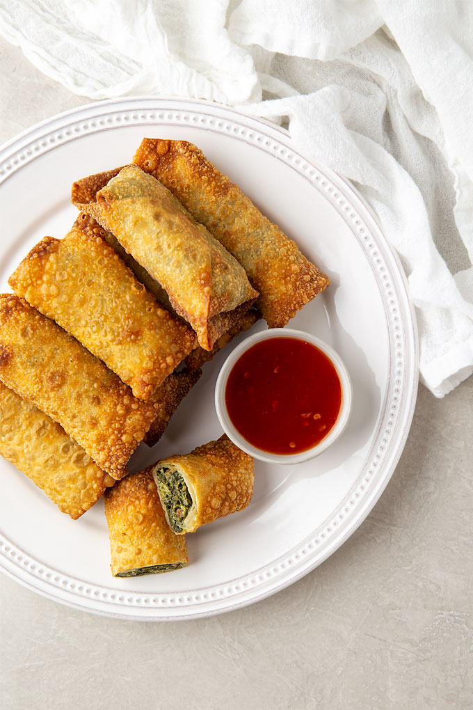 Collard green egg rolls on a round white plate with a small bowl of chili sauce