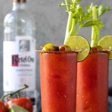 Two bloody mary cocktails with garnishes. A bottle of vodka, tomatoes and a lime is in the background