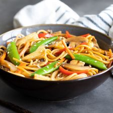 Vegetable Lo Mein in a blue bowl beside chopsticks. A striped kitchen towel is in the background.