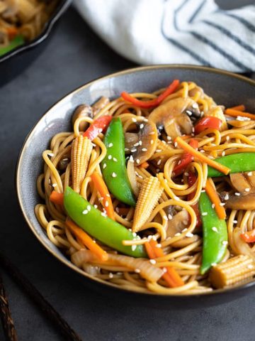 Vegetable lo mein in a blue bowl beside chopsticks. A striped kitchen towel and skillet is in the background.