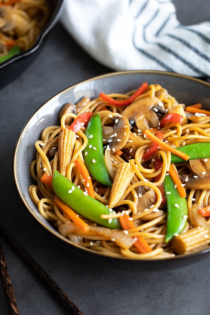 Vegetable lo mein in a blue bowl beside chopsticks. A striped kitchen towel and skillet is in the background.