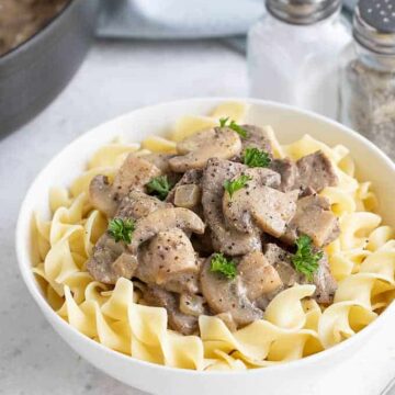 Beef stroganoff over egg noodles in a white bowl. Salt and pepper shakers and a skillet is in the background.