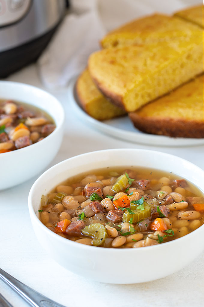 Two white bowls of ham and bean soup with a plate of cornbread in the background