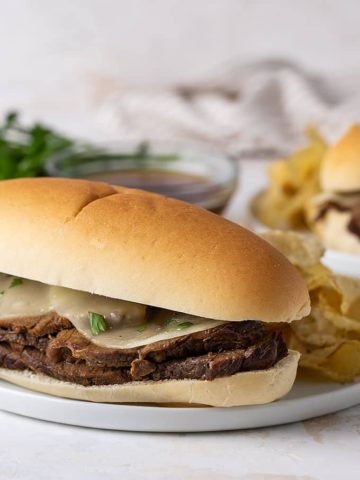 A French dip sandwich with melted provolone on a white plate. A bowl of au jus is in the background