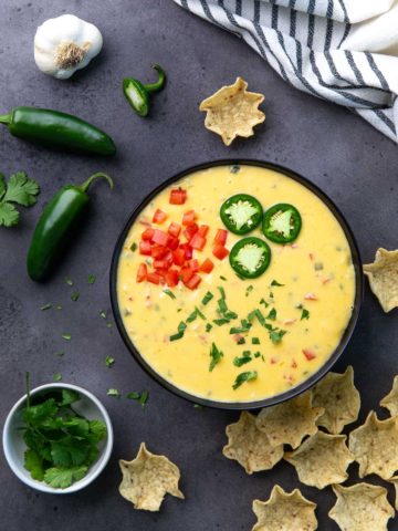 Overhead view of queso dip in a black bowl topped with cilantro, tomatoes and sliced jalapeno.