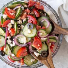 Overhead view of Greek cucumber tomato salad in a glass bowl with wooden salad servers in the bowl.