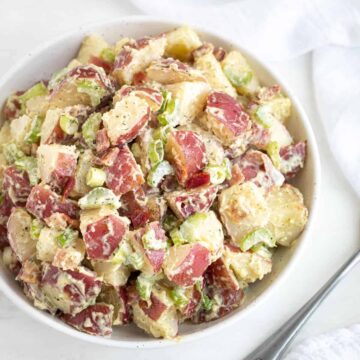 Overhead view of potato salad in a white bowl by a serving spoon and a white napkin.