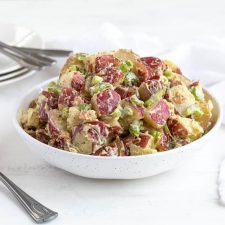Front view of a bowl of potato salad. Serving plates and forks are in the background.