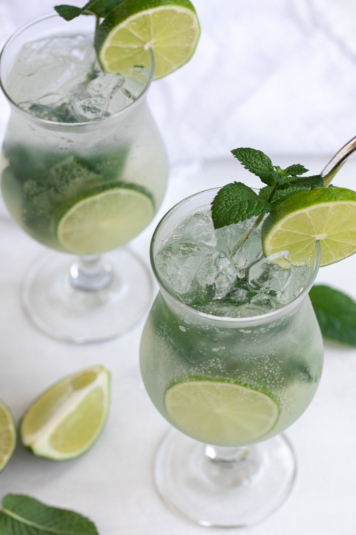 Angled overhead view of two mojito cocktails on a white surface.