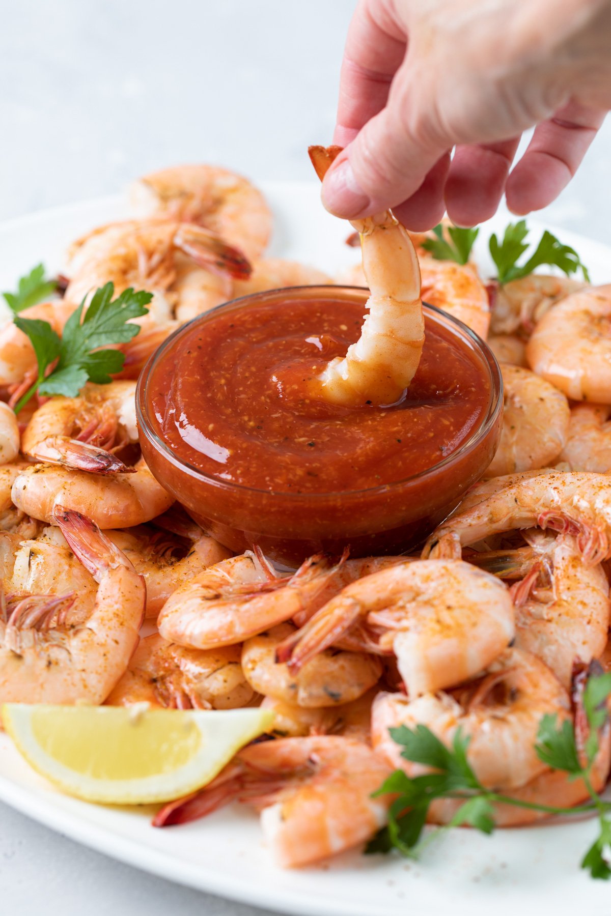 Dipping a shrimp into a bowl of cocktail sauce on a white platter with shrimp.