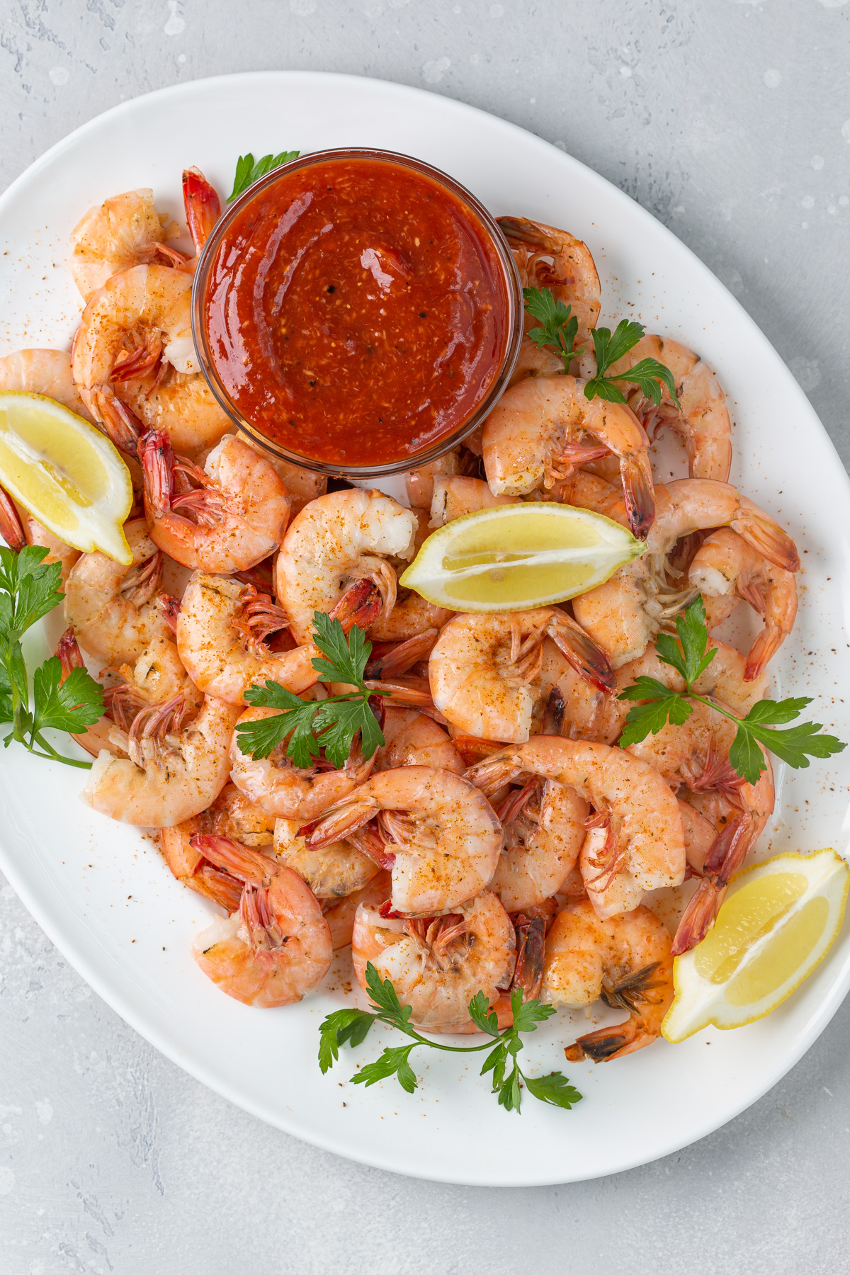 Overhead view of a white platter of steamed shrimp with a bowl of sauce and lemon wedges.