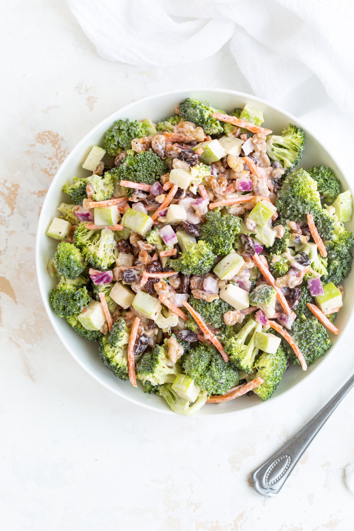 Overhead view of creamy broccoli salad in a white bowl beside a spoon.