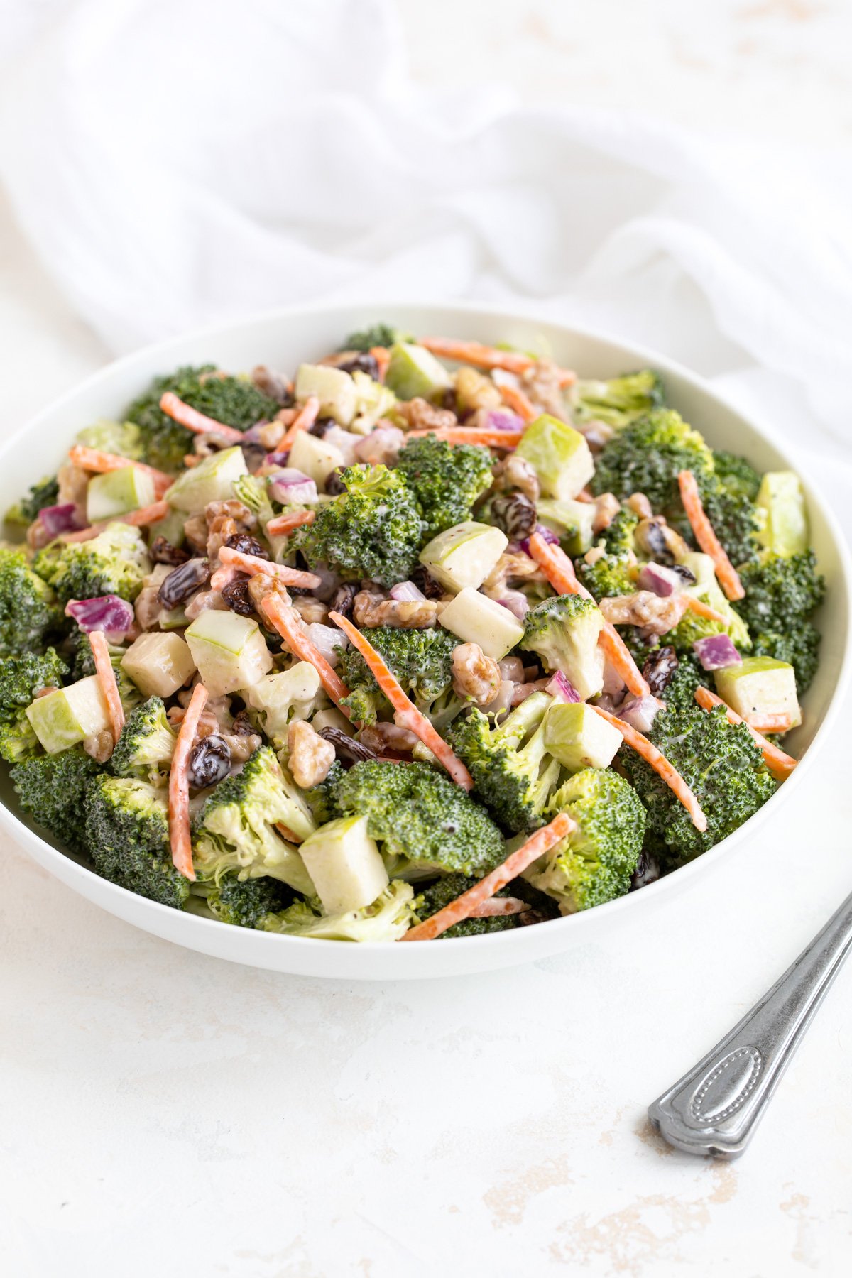 Creamy broccoli salad in a white bowl beside a white napkin and a spoon.