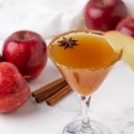 A martini with red apples and cinnamon sticks in the background.