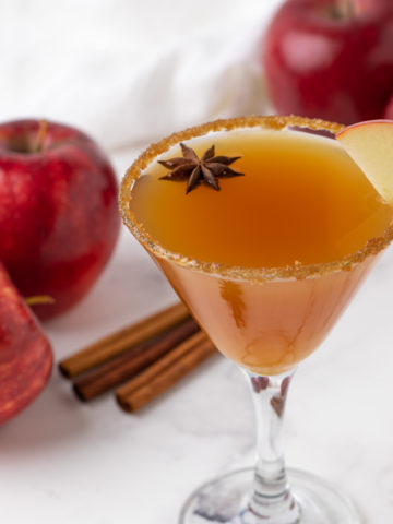 A martini with red apples and cinnamon sticks in the background.