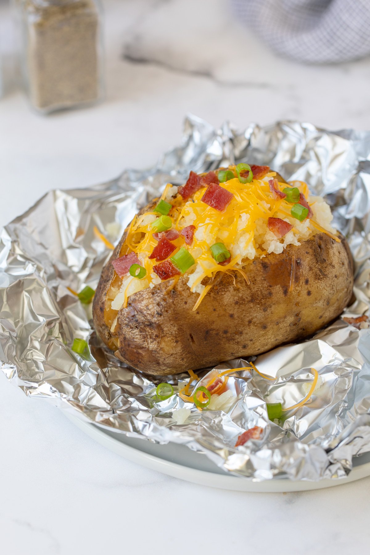 A baked potato topped with cheese, bacon and green onion on a piece of aluminum foil.