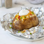 A slow cooked russet potato with toppings on top of a piece of aluminum foil.