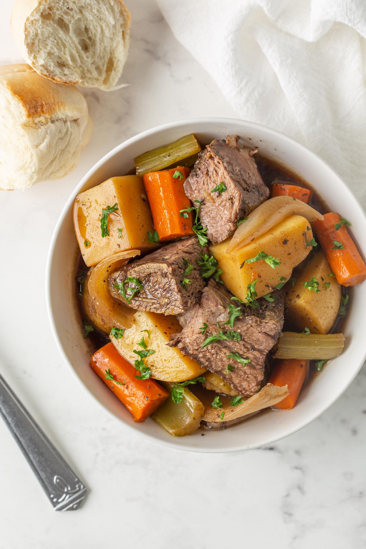 Overhead view of a white bowl of pot roast and vegetables by a bread roll.